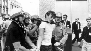 Black and white image of three firefighters assisting an injured man away from a scene, with bystanders and a high fence in the background.