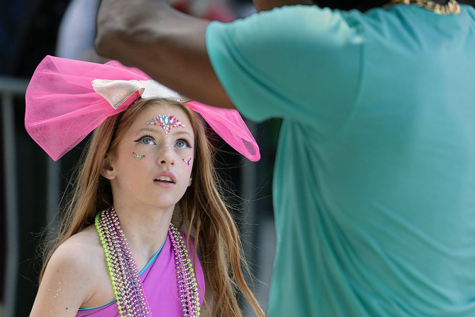 A young girl with a large pink bow and colorful face paint looks up at a person in a turquoise shirt during an outdoor event.