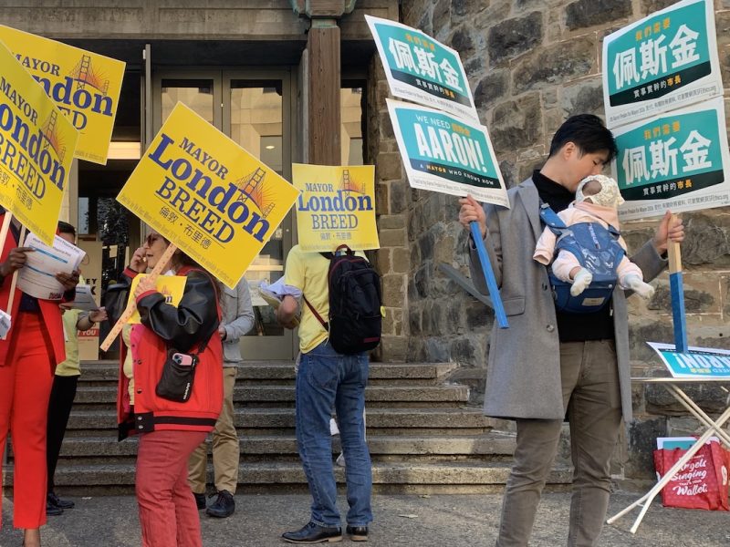 People holding campaign signs supporting Mayor London Breed and another candidate, standing outside a stone building. One person is holding a baby. Signs are written in both English and Chinese.