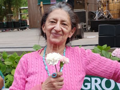 Denhi Donis, the flower lady, in a pink sweater smiles while holding white and pink flowers at an outdoor event.