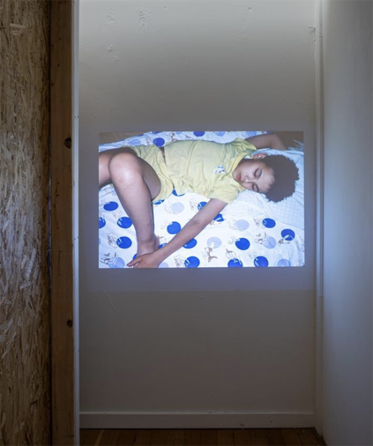 A photograph of a child sleeping on a blue and white patterned bed is projected on a white wall in a narrow space.