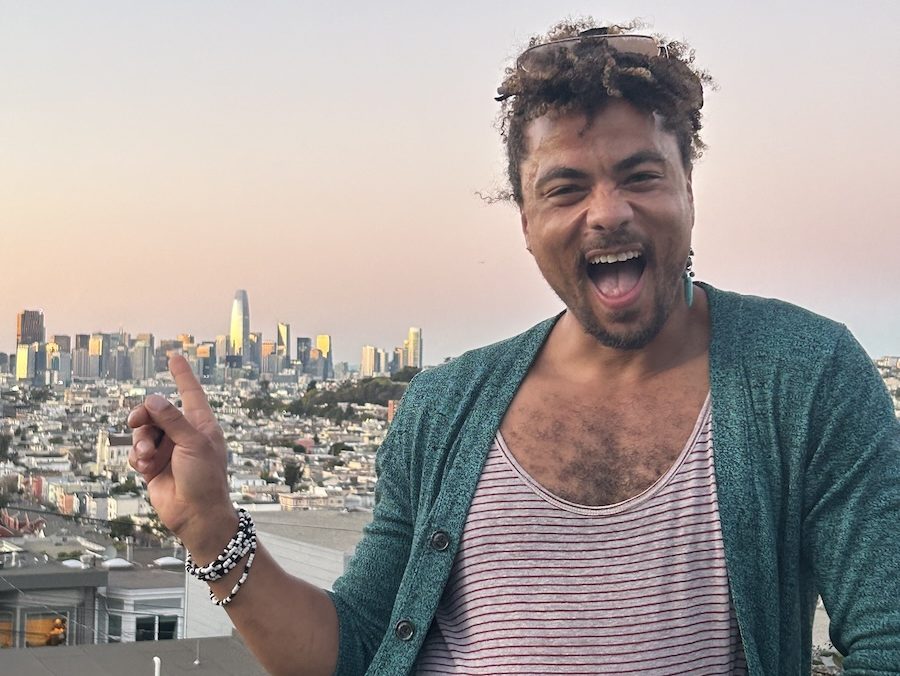 Person with curly hair, wearing a teal cardigan and striped shirt, smiles widely and points towards a city skyline at sunset.