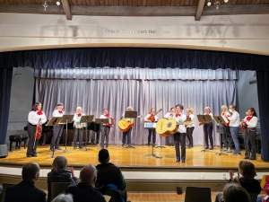 A group of musicians performs on stage in front of an audience. They play string instruments and are dressed in white shirts with red accessories. The backdrop is a blue curtain.