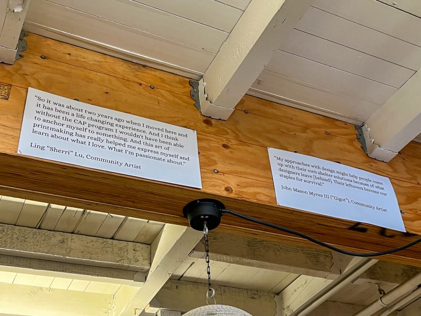Two quotes from community artists are displayed on the wooden beam of a ceiling. The space has visible structural elements and a light fixture hanging below one of the beams.