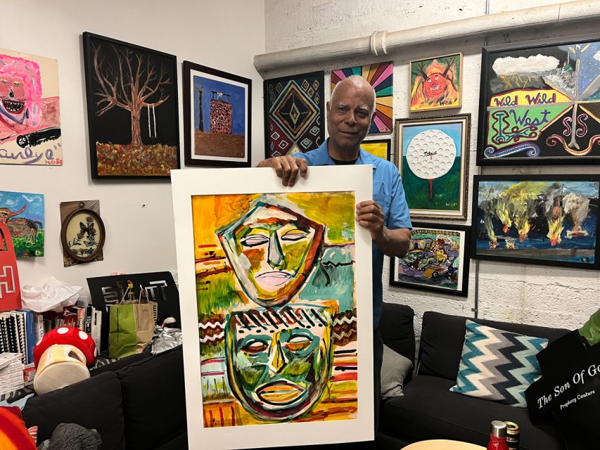 A person is holding a colorful painting featuring two abstract faces. The room has multiple artworks displayed on the walls and some items on a couch and a table.