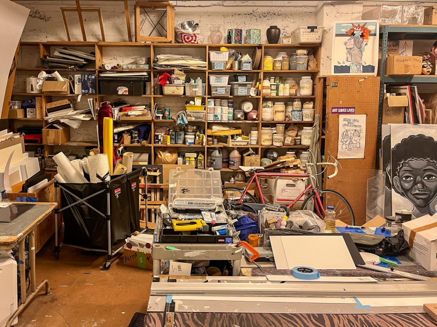 A cluttered art studio with shelves full of various supplies, canvases, jars, and tools. A bicycle is positioned in the center. A large drawing of a face is propped up on the right.