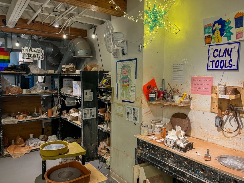 A pottery studio with clay tools, workstations, shelves with pottery pieces, a "CLAY TOOLS" sign, and a wall adorned with various art pieces. A fairy light star decoration hangs from the ceiling.