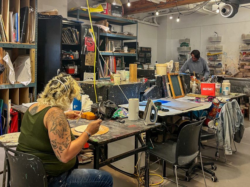 Two people work in an art studio filled with supplies. One person paints at a cluttered table with art tools, while the other stands at the back near shelves. Various art materials are visible.
