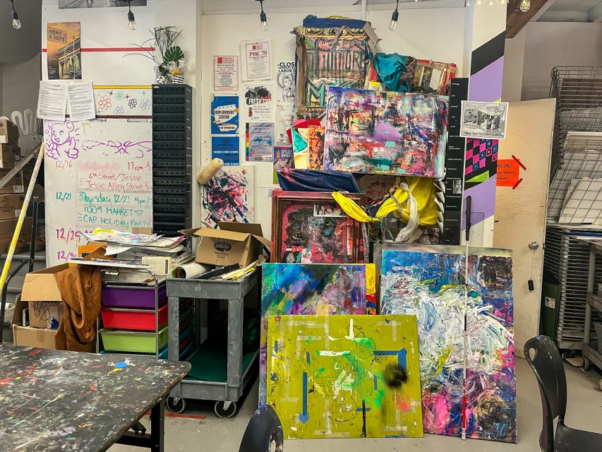 A cluttered art studio with colorful abstract paintings, storage carts, and various art supplies scattered around on shelves and tables. Papers are pinned on the walls, and some furniture is overturned.