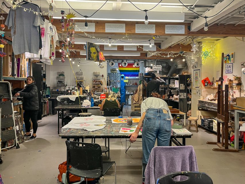 Three people are working in a cluttered art studio filled with various supplies, artworks, and tools. The central person is painting at a table, while another is organizing shelves. The space is well-lit.