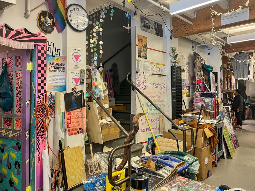 A cluttered art studio brightly decorated with a variety of colorful artwork, posters, and signs. A clock shows the time as 3:05. Shelves and tables are filled with art supplies and materials.