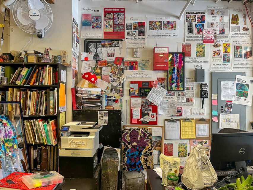 A cluttered office space with walls covered in colorful posters, flyers, and artwork. There are shelves filled with books on the left and a desk with various items on the right.