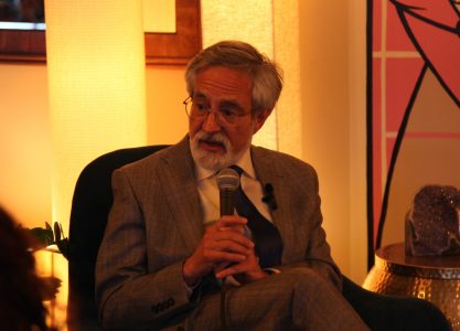 An elderly man with a beard and glasses, identified as Aaron Peskin, dressed in a suit, speaking into a microphone while seated indoors.