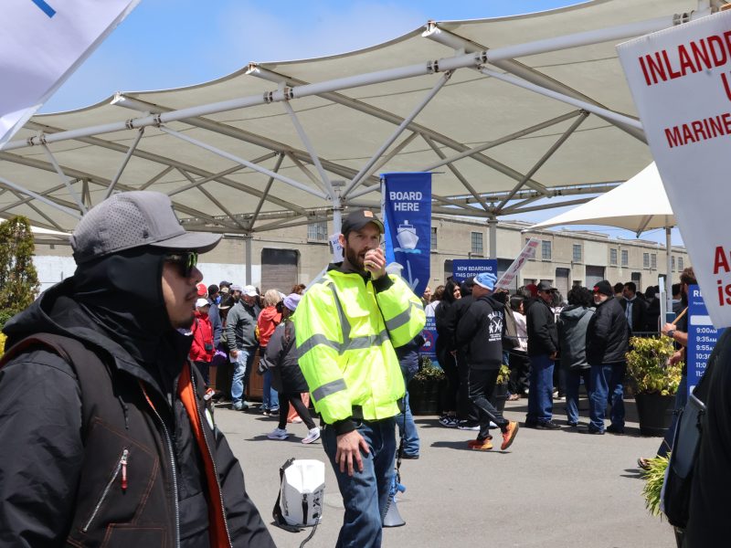 A man in a bright yellow jacket speaks into a handheld radio while standing in a crowded outdoor area. Various signs and people are visible in the background.