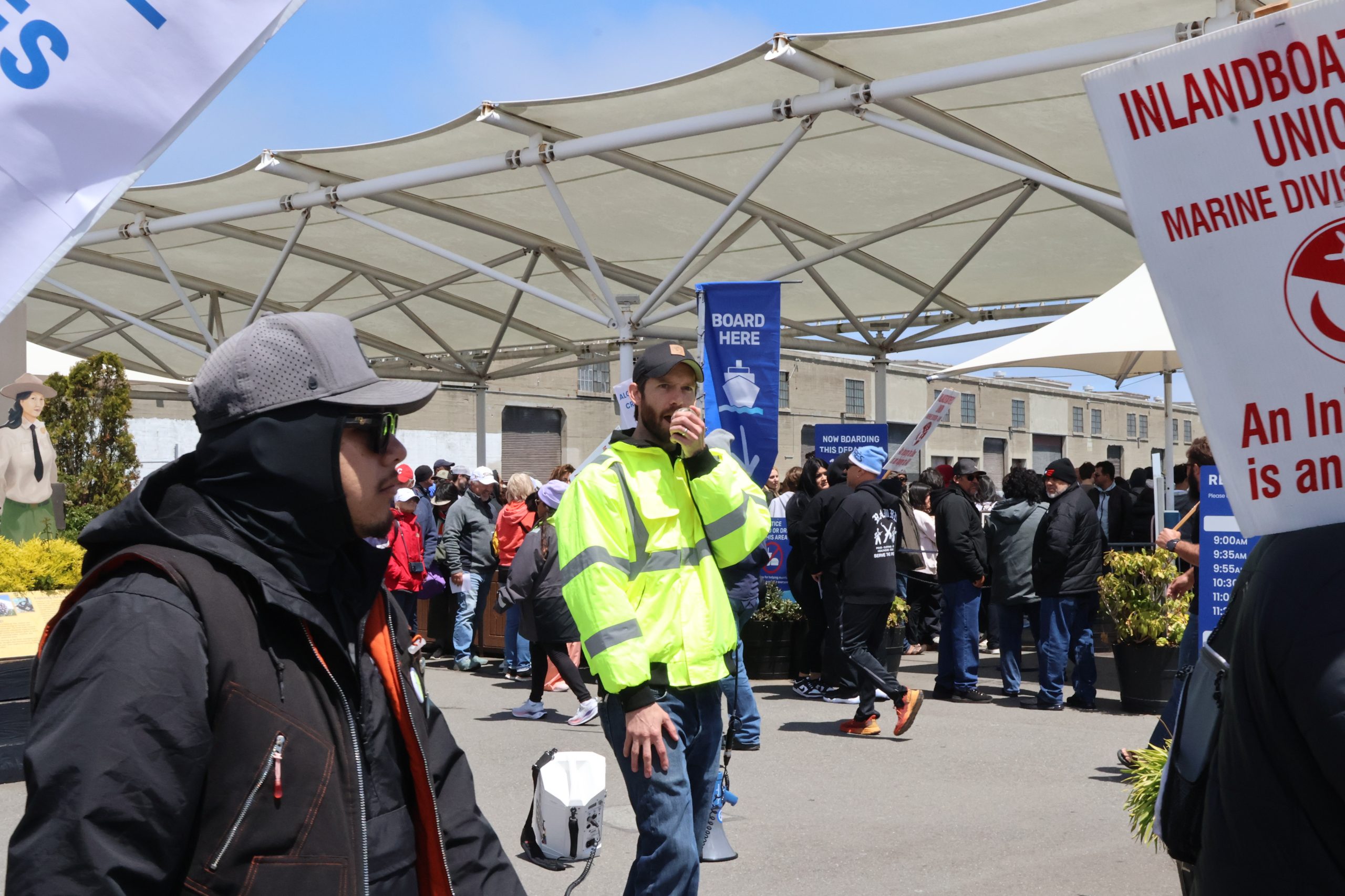 A man in a bright yellow jacket speaks into a handheld radio while standing in a crowded outdoor area. Various signs and people are visible in the background.