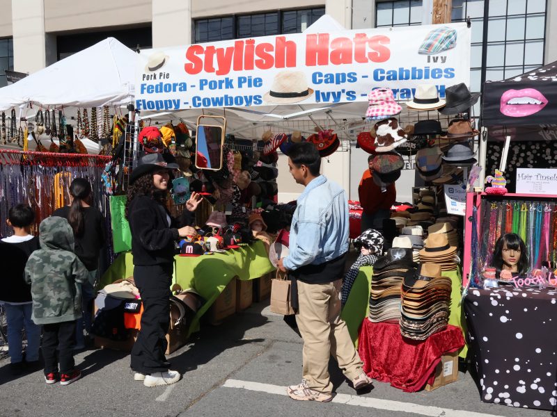 People shopping at an outdoor market stall selling various styles of hats, with a sign overhead that reads "Stylish Hats". Various colorful hats and accessories are displayed.