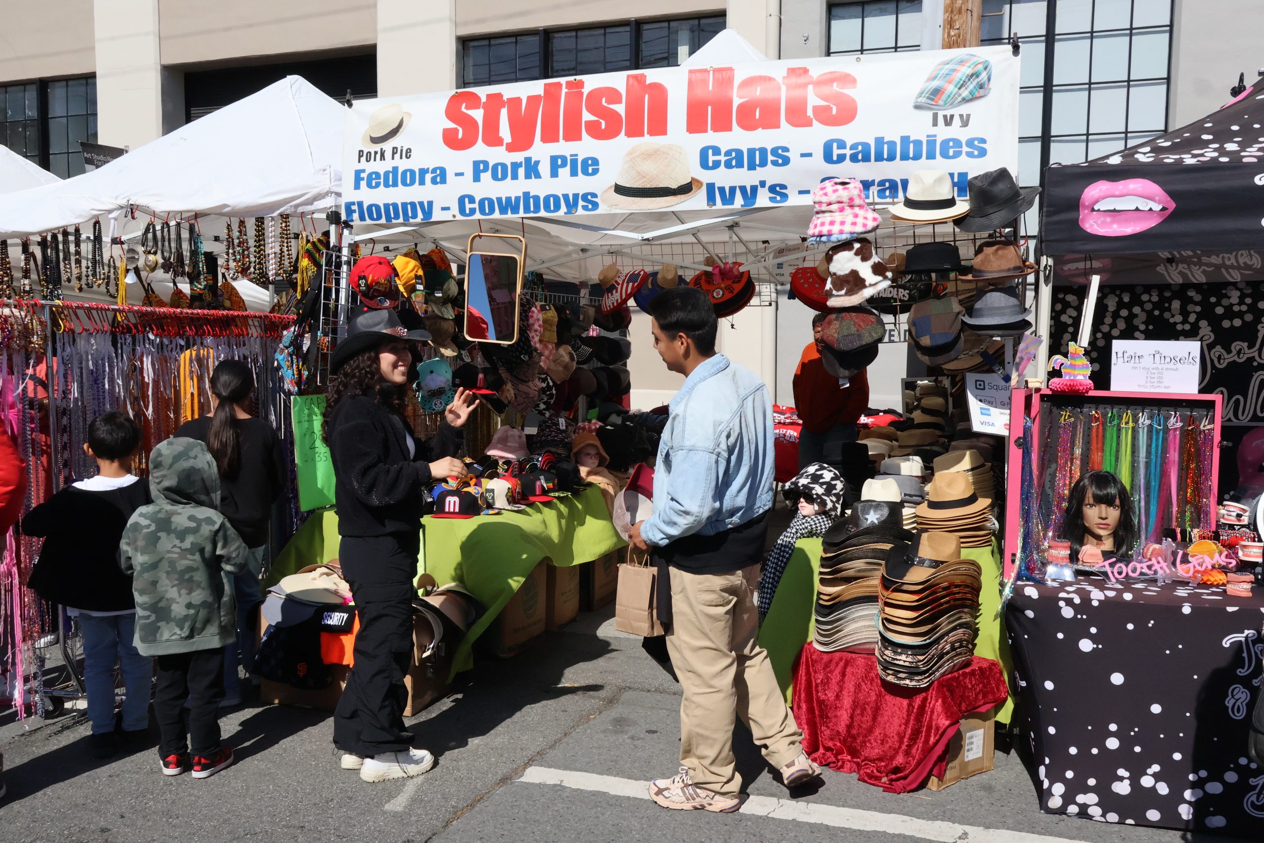People shopping at an outdoor market stall selling various styles of hats, with a sign overhead that reads "Stylish Hats". Various colorful hats and accessories are displayed.
