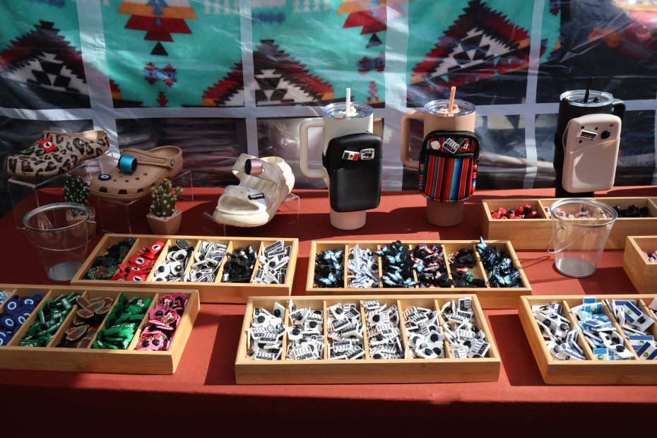 A table display features various small decorative items organized in wooden trays, along with decorated clogs, drink holders, and assorted ornaments. The background has a colorful geometric-patterned fabric.