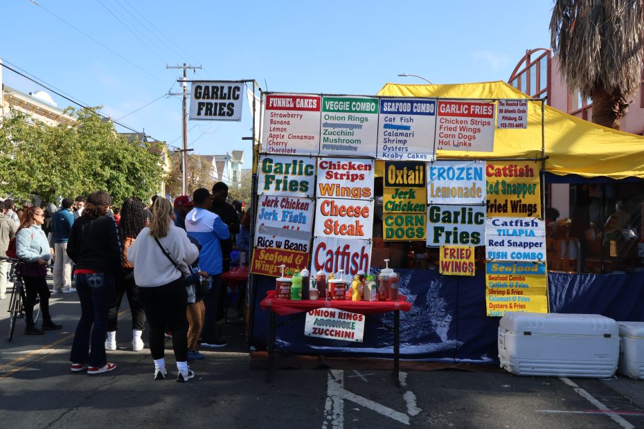 People standing in line at a food stall offering various fried foods, garlic fries, and drinks at an outdoor event. The menu includes options like catfish, oxtail, chicken strips, and cheese steak.