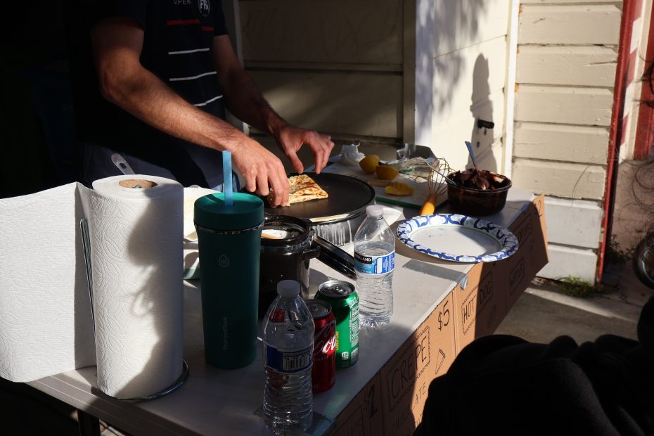 A person is preparing food outside on a table. Various items, including paper towels, a water bottle, soda cans, and plates, are on the table.