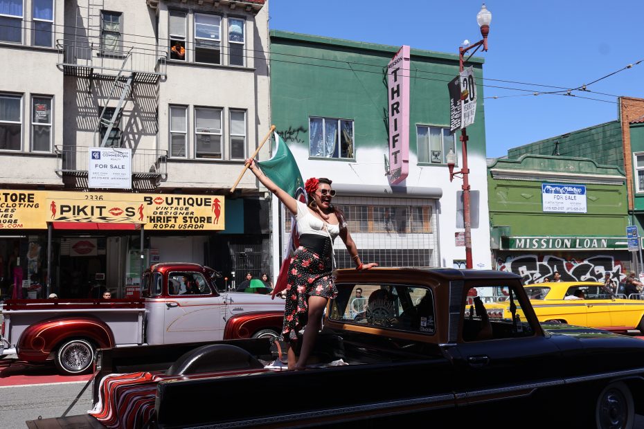 A woman standing and waving a flag on the back of a pickup truck in a lively street scene featuring several classic cars and storefronts in the background. At Carnaval. 