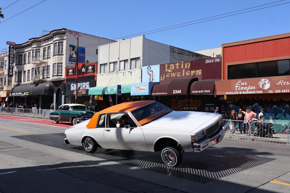 A white car with an orange roof is performing a hydraulic lift on a street in front of various storefronts, including shops and restaurants. Another car is parked in the background, and people are watching. At Carnaval. 