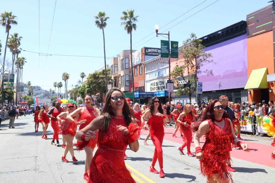 A group of people in red costumes dances in a street parade, surrounded by spectators. The area is lined with palm trees and vibrant buildings. at Carnaval. 
