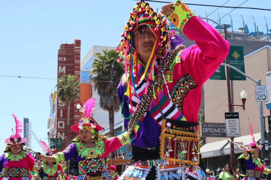 Colorful street festival with participants in traditional, vibrant attire. A person in a bright pink outfit and decorated hat is centered. Buildings, signs, and the sky are visible in the background. At Carnaval. 