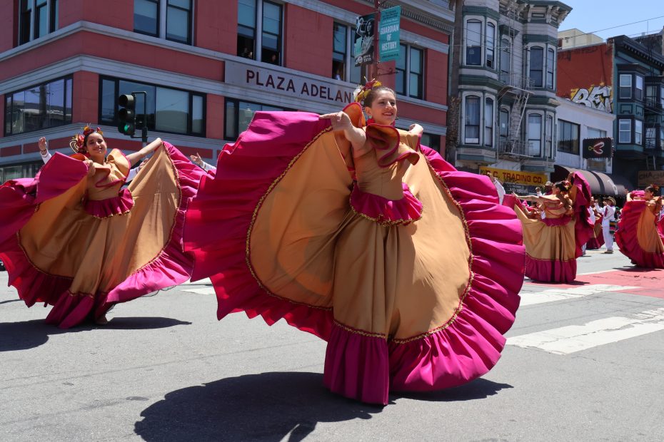 Female dancers in colorful dresses perform in the street during a parade in front of Plaza Adelante, with buildings and spectators in the background. At Carnaval. 