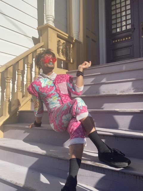 Person in colorful clothing and red sunglasses sits on outdoor steps, leaning back with one hand raised. They are wearing black shoes and large earrings, with a purple door in the background.