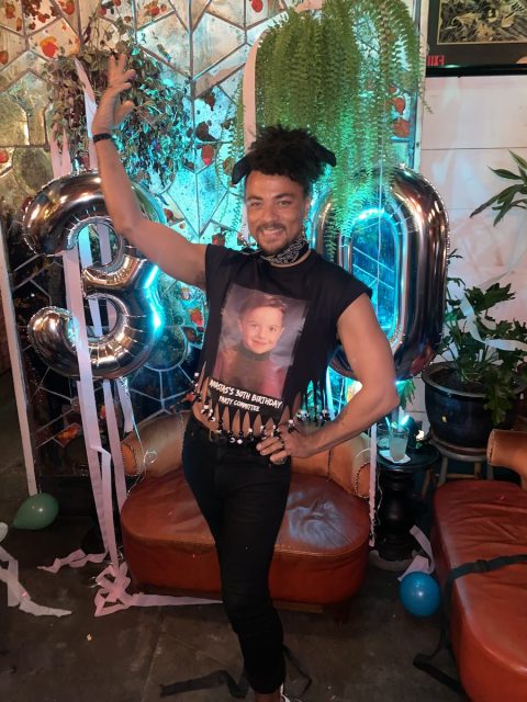 A person poses with an arm raised in front of large metallic "30" balloons, wearing a sleeveless shirt with a childhood photo printed on it. Decorations and plants are visible in the background.