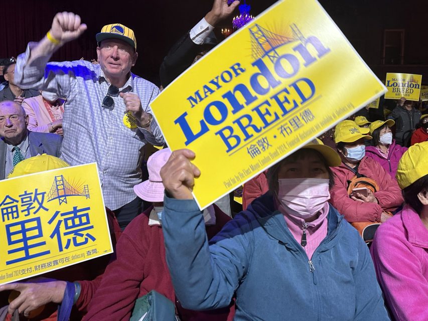 A group of people hold up yellow signs reading "Mayor London Breed" at an indoor event. Some attendees wear hats and masks.