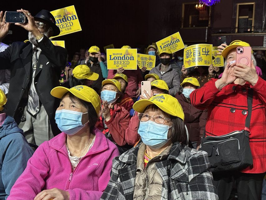 A seated crowd with many people wearing yellow hats and masks hold up yellow signs that say "London Breed" while some use their phones to take photos and videos.