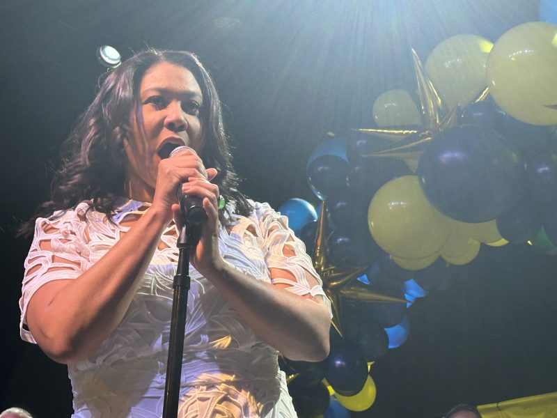 A woman in a white dress speaks into a microphone on stage, with a backdrop of blue and yellow balloons under bright overhead lighting.