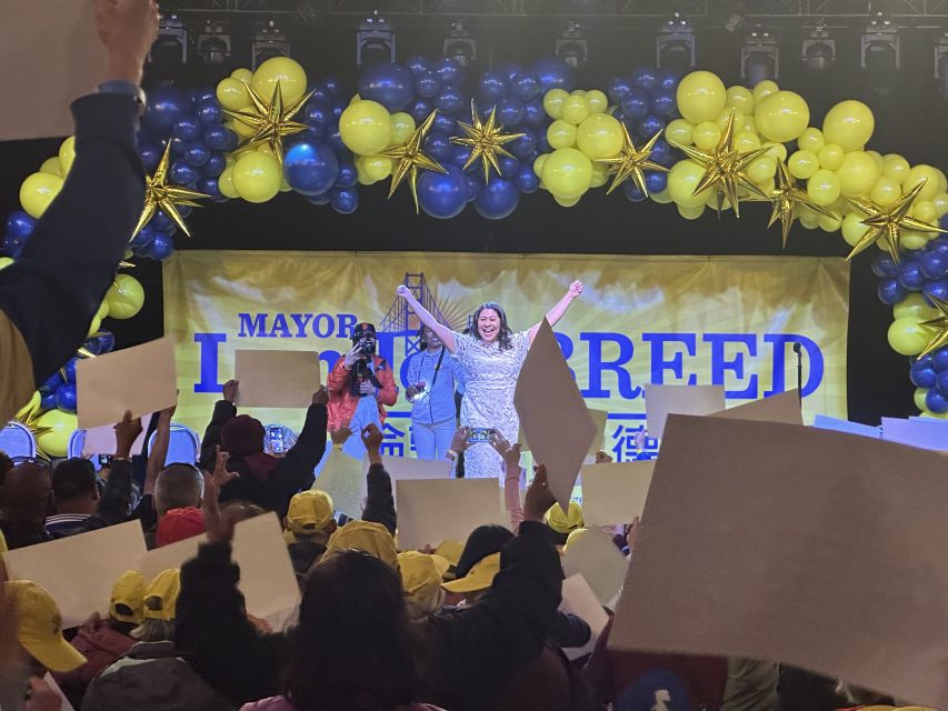 A person stands on stage with arms raised, surrounded by yellow and blue balloons, in front of a sign that says "Mayor." A crowd holding signs and wearing yellow hats cheers in the foreground.