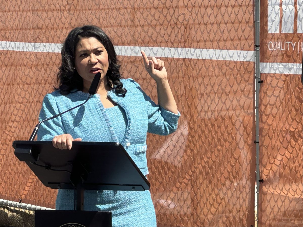 London Breed, in a blue pantsuit, speaks in front of a podium and points