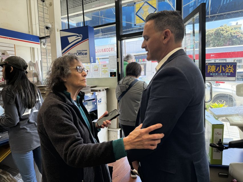 An older woman talks with a man in suit, holding his arm