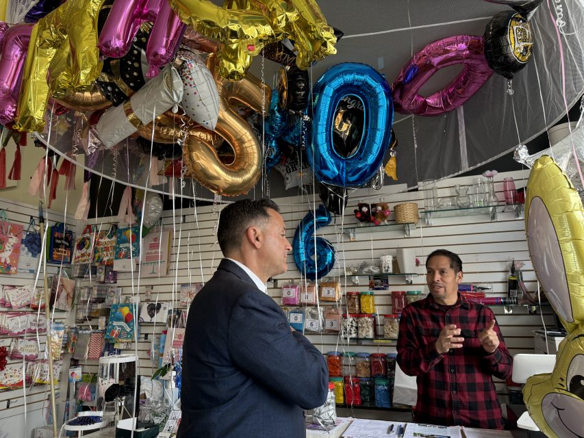 Two men in conversation at a party supply store, surrounded by colorful balloons, decorations, and merchandise displays.