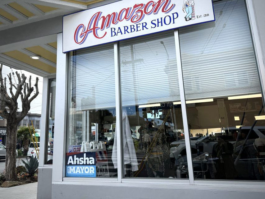 A storefront with a sign reading "Amazon Barber Shop." The window displays a sign that says "Ahsha for Mayor." Inside, a haircut is in progress.