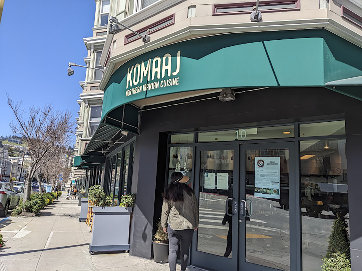 A person wearing a hat stands in front of Komaj, a Northern Iranian cuisine restaurant, located on a sunny city street.