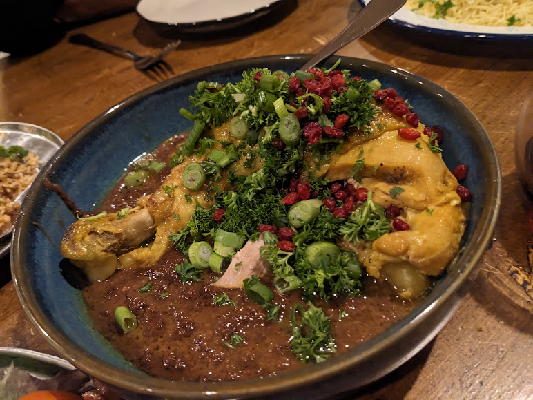 A bowl of stewed chicken breast garnished with chopped green onions, parsley, and pomegranate seeds, served in a rich, dark sauce.