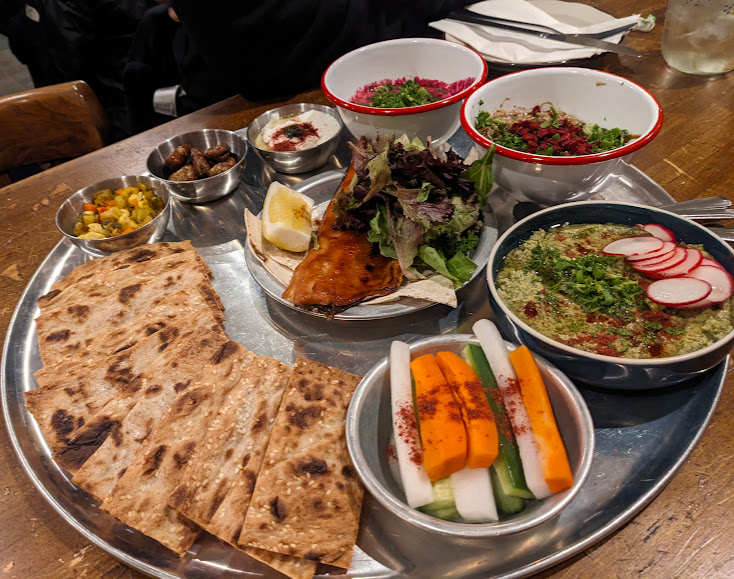 A large metal tray holds various dishes including flatbreads, hummus, vegetable sticks, salad, and other Mediterranean cuisine items, arranged neatly.