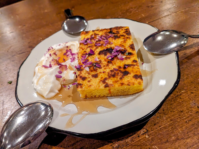A slice of baked dessert served on a plate, garnished with edible flowers, syrup, and a dollop of cream. Three spoons surround the plate, ideal for sharing. The dessert sits on a wooden table.