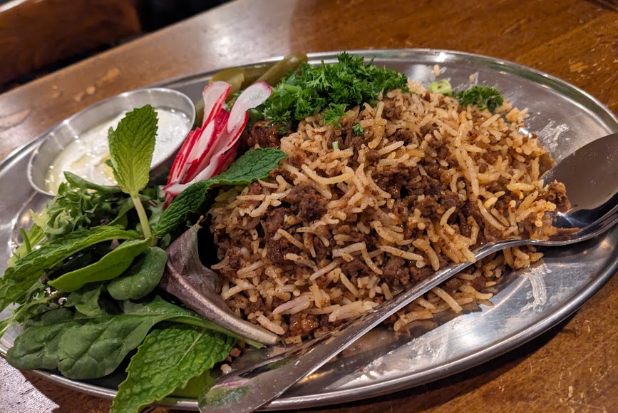 A metal plate with a serving of seasoned rice mixed with minced meat, accompanied by fresh herbs, sliced radishes, and a dollop of yogurt on the side.