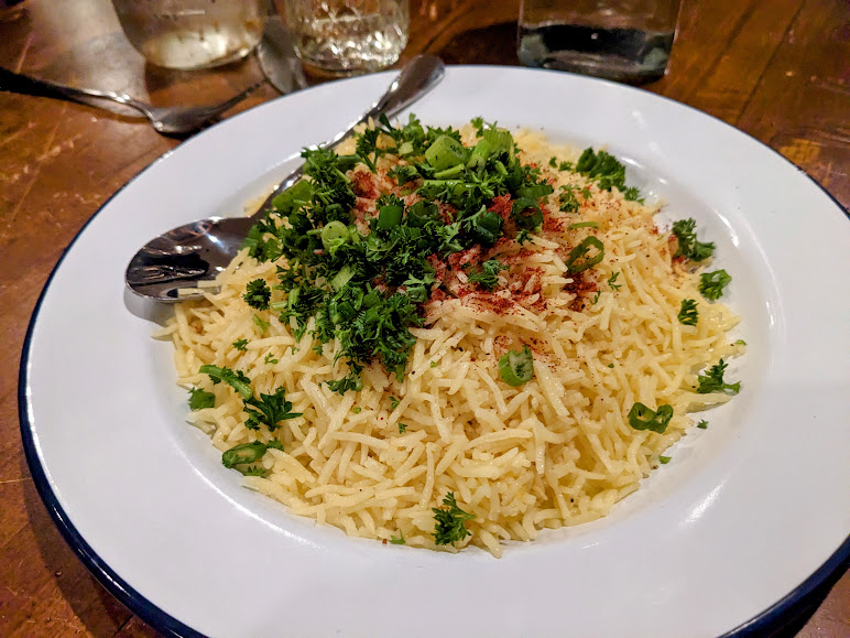 A plate of seasoned rice garnished with chopped green herbs and spices, served with two metal spoons.