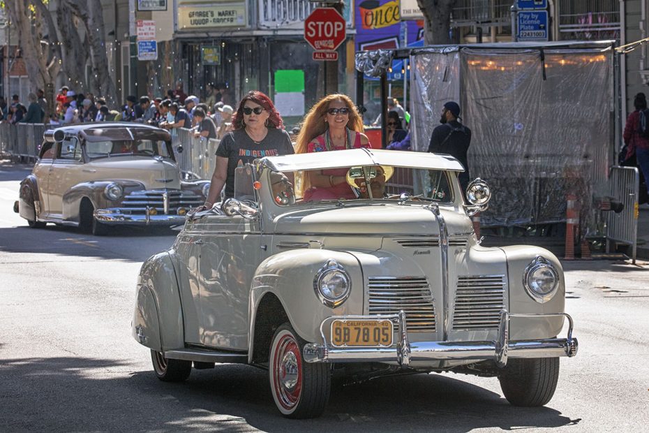 Two women drive a vintage convertible car in a parade, followed by another classic car. A crowd lines the street under a stop sign and shop signs in the background.