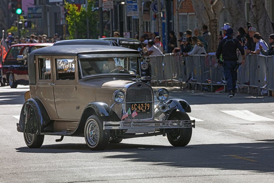 A vintage car with a "GC-4279" license plate drives down a street lined with spectators behind barricades.