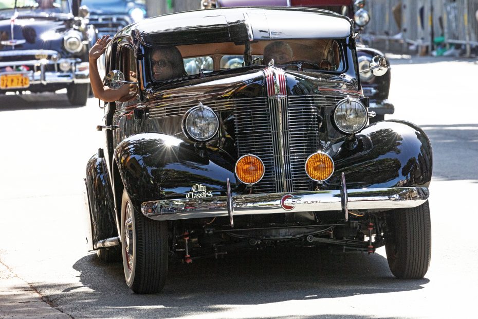 A black vintage car with two people inside drives down a sunny street. The passenger waves their hand out of the window. Other classic cars are seen in the background.