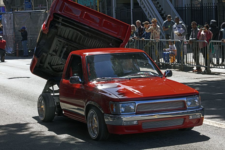 A red pickup truck with its bed lifted vertically is parked on a street. People are standing behind a barricade in the background.