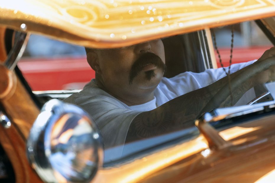 A man with a mustache is seen driving a vintage car with a reflective bronze interior. He is partially obscured by the rearview mirror and the car's windshield frame.